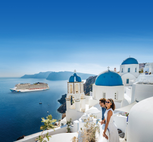 Paar blickt von den weißen Häusern mit blauen Kuppeln in Santorini auf ein Kreuzfahrtschiff im tiefblauen Meer.
