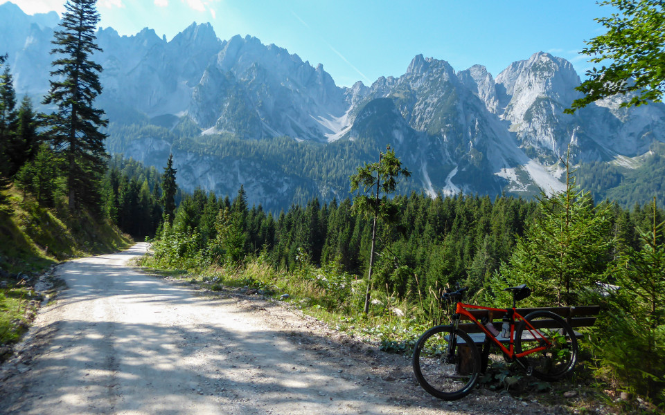 Ein orangefarbenes Mountainbike lehnt an einer Bank neben einer Schotterstraße in den Bergen mit Blick auf die hohen Alpen in der Region Gosau, Österreich.