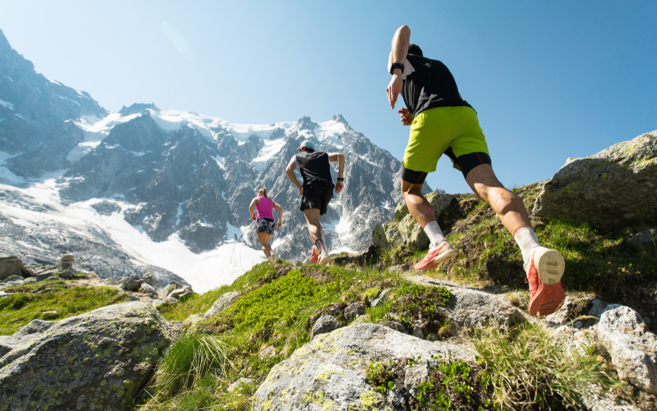 Drei Trailrunner, zwei Männer und eine Frau, laufen an einem heißen, hellen Sommertag einen steilen Pfad in den Bergen der Alpen hinauf.  