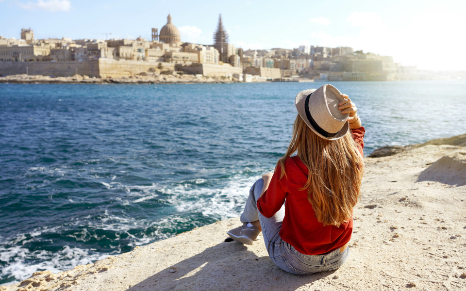 Schöne junge Frau mit Hut sitzt auf einem Stein am Meer und blickt auf einen atemberaubenden Panoramablick auf die Stadt Valletta auf Malta