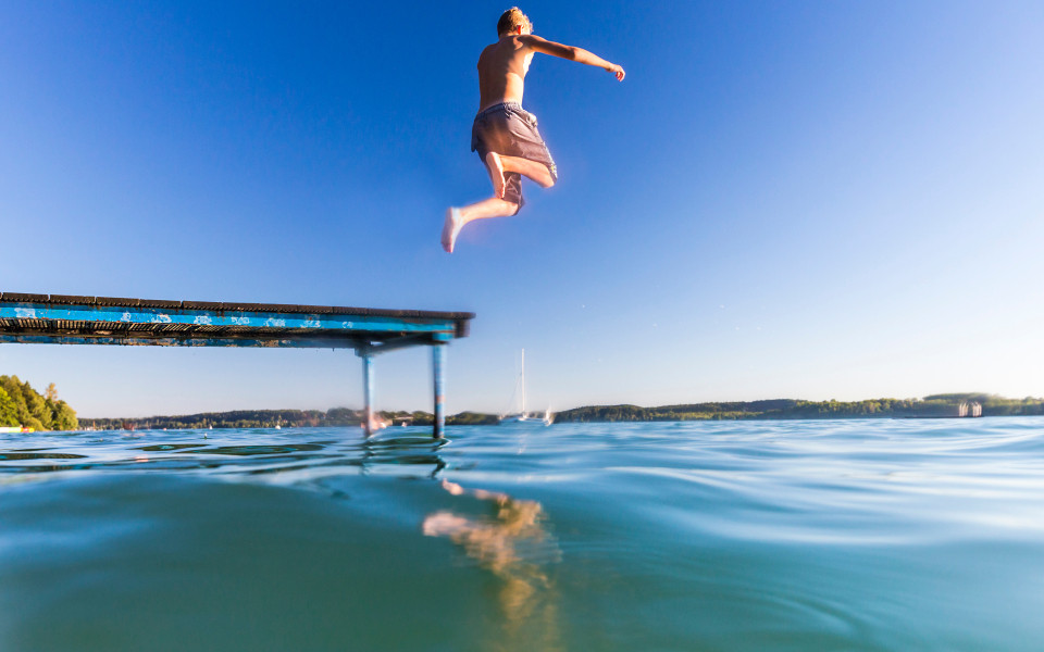 Junge in Badehose hüpft vom Steg in den Chiemsee mit Alpenblick im Hintergrund