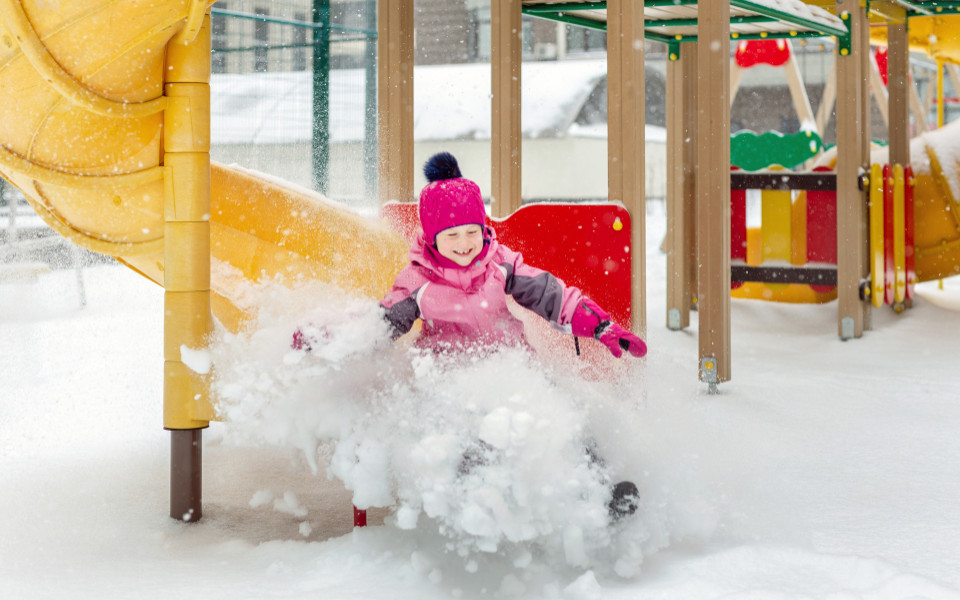 Kleines Mädchen mit pinker Mütze und Winterjacke, das sich im Winter auf dem Spielplatz vergnügt.