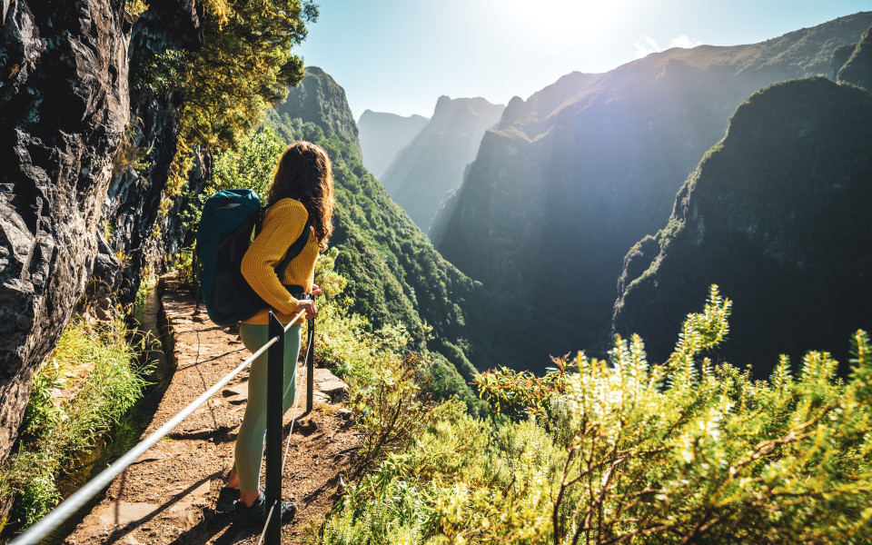 Eine Rucksacktouristin genießt die malerische Aussicht von unterhalb einer großen Felswand entlang eines Wasserlaufs an einer steilen Klippe im Regenwald von Madeira.