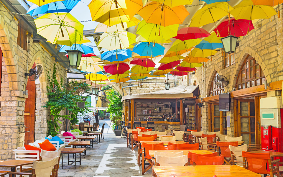 Bunte Regenschirme hängen über einer schmalen, mediterranen Gasse mit Natursteinfassaden und kleinen Café-Tischen in Limassol.